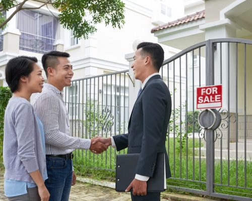 Cheerful Vietnamese real estate agent meeting new client in front of house for sale